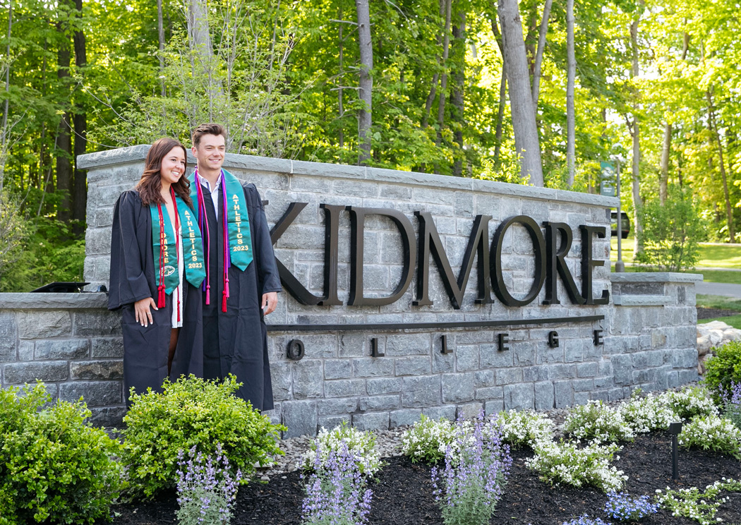 two college graduates post in front of a campus entrance sign that says С